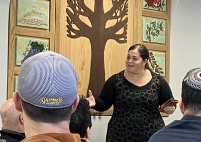 A woman gesturing while speaking to a group of seated people in a room with a wooden tree artwork on the wall.