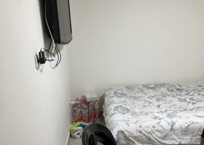 A corner of a sparse room with a mounted tv, an unmade bed, a black chair, and bottles on the floor.