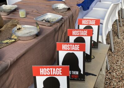 A banquet table outdoors with empty plates and signs reading "hostage - bring them home now!" indicating a protest or awareness event for hostages.