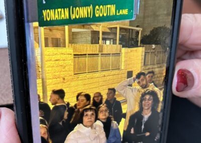 People looking up, with a street sign reading "yonatan (jonny) goutin lane" in the foreground, displayed on a smartphone screen.