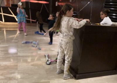 A child in pajamas stands near a reception desk while other children play in the background of a dimly lit lobby area.