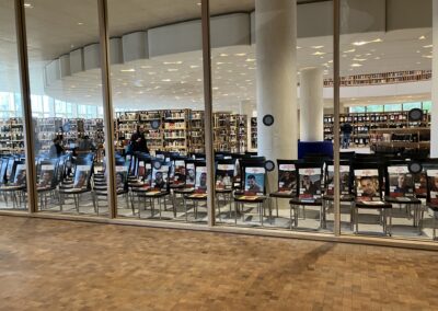 A public library interior with bookshelves and a row of promotional standees visible through glass windows.