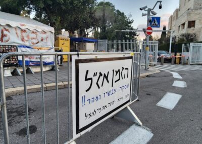 Barricade with a sign in hebrew blocking a street, with tents and buildings in the background.